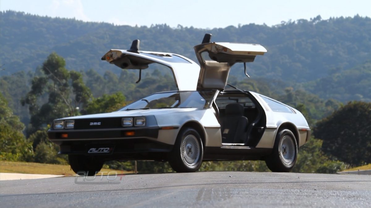 DeLorean DMC-12 parado no asfalto com as portas verticais abertas com montanha e árvores ao fundo