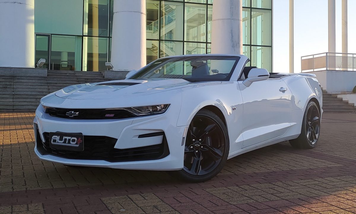 Entre os modelos conversíveis à venda em nosso mercado está o Chevrolet Camaro SS Conversível branco parado de frente com prédio ao fundo