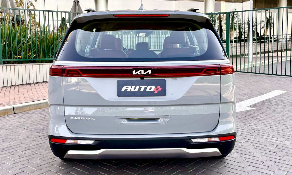 A Kia sofreu um processo de mudança de identidade de marca e a  Carnival está parada de traseira com portão e plantas ao fundo [Auto+ / João Brigato]