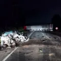 Acidente de trânsito a noite com carro prata destruído e caminhão vermelho bloqueando a via