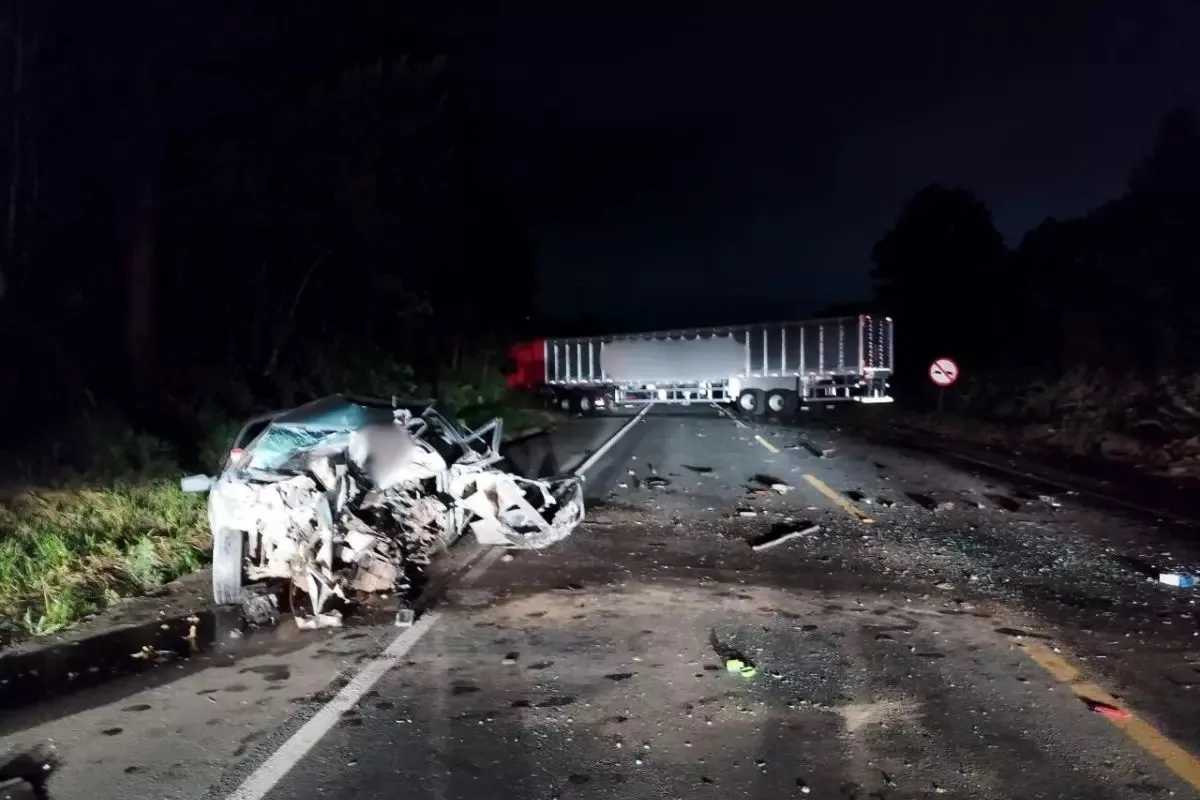 Acidente de trânsito a noite com carro prata destruído e caminhão vermelho bloqueando a via