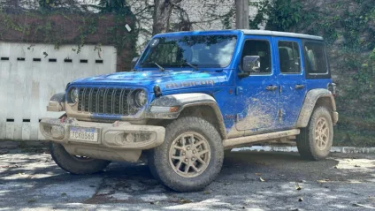 Jeep Wrangler Rubicon azul parado de frente com árvore e portão branco ao fundo