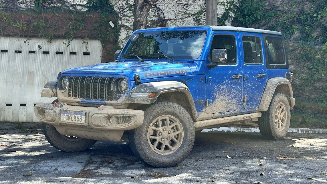 Jeep Wrangler Rubicon azul parado de frente com árvore e portão branco ao fundo