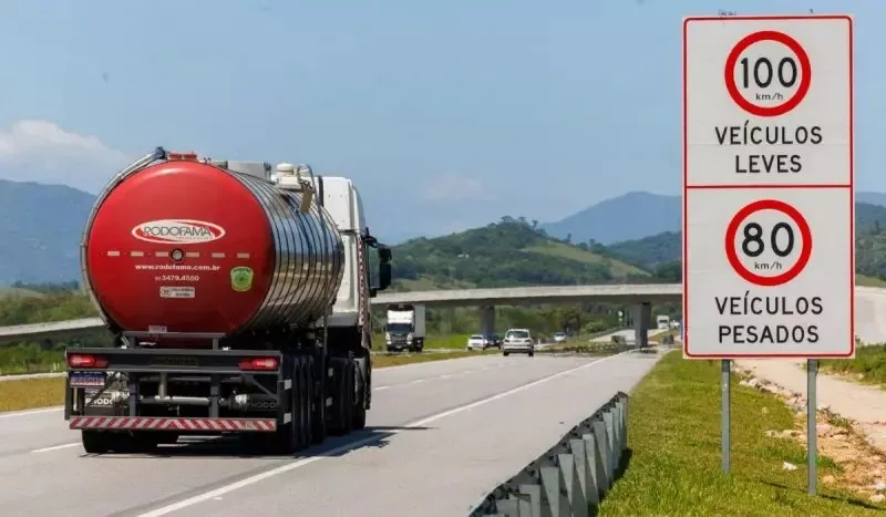 Caminhão visto de trás, andando em uma estrada, com carros ao redor e uma placa com limites de velocidade