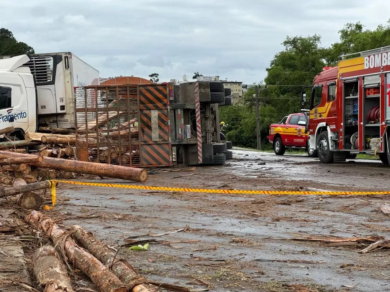 Caminhão tombado após acidente, com madeira espalhada pela pista e ambulâncias ao redor