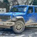 Jeep Wrangler Rubicon azul parado de frente com árvore e portão branco ao fundo