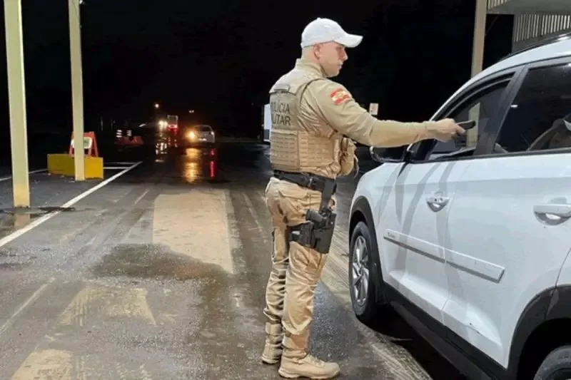 Agente da polícia rodoviária federal entregando documento para motorista que está a bordo de um carro branco com fundo escurecido