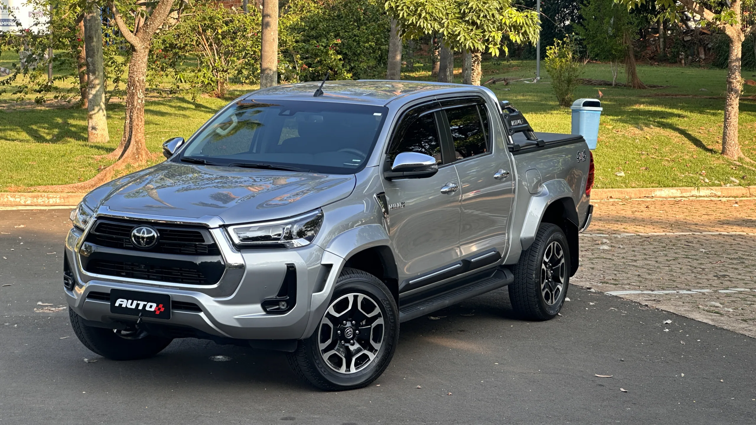 Toyota Hilux SRX Plus prata parada de levemente de lado em um estacionamento com uma floresta ao fundo