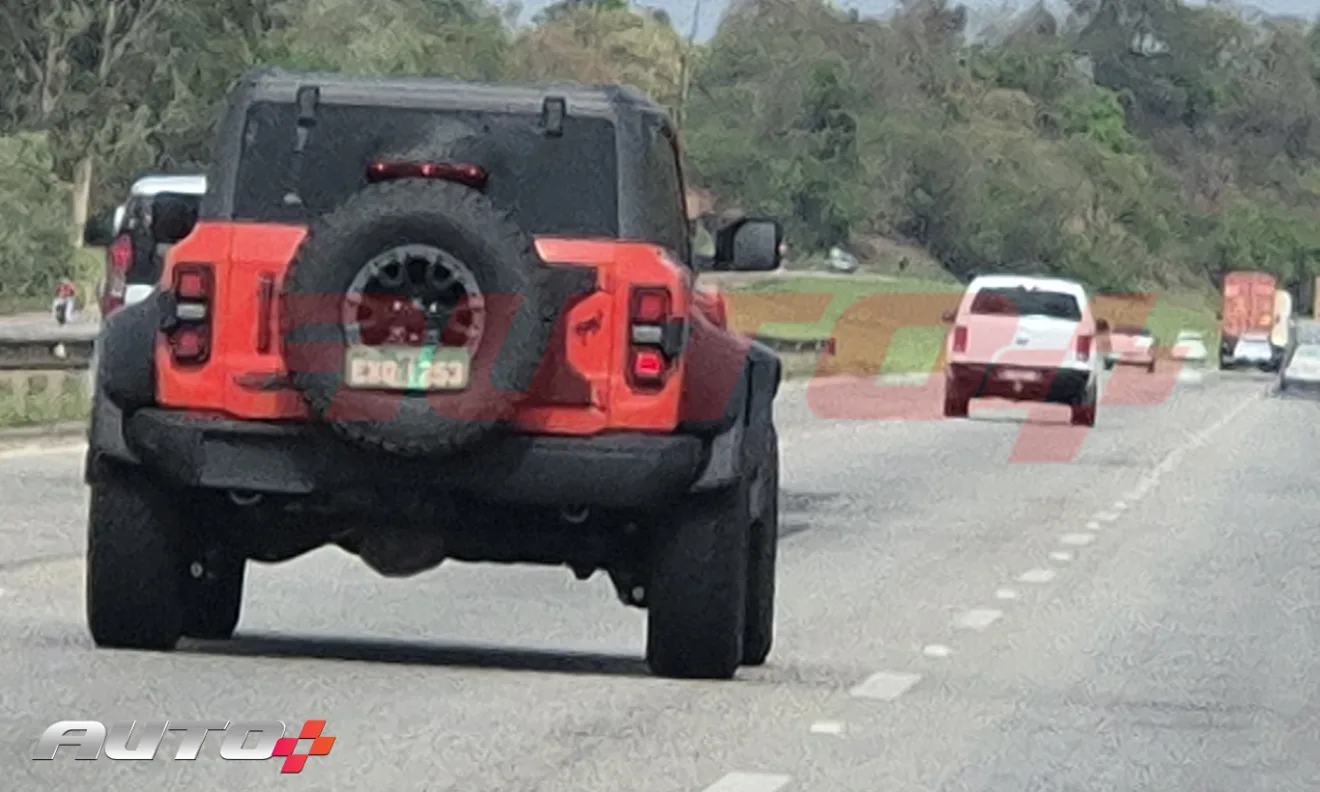 Ford Bronco Raptor laranja com partes pretas, visto de trás em uma rodovia e atrás de uma caminhonete branca