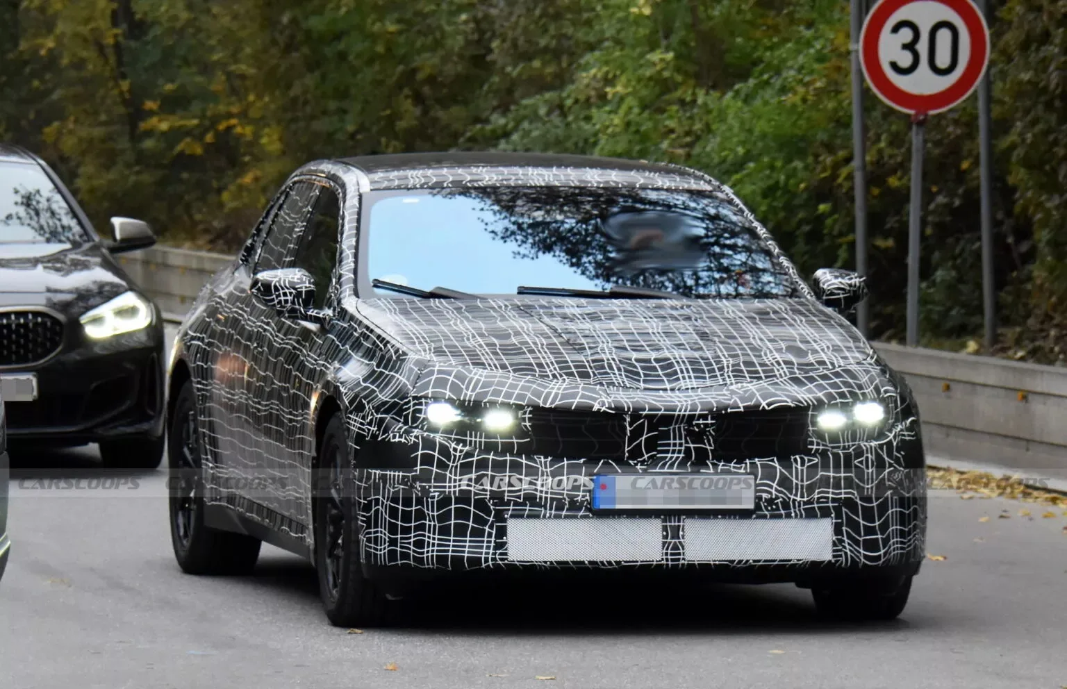 Protótipo do BMW i3 sedã com pintura escurecida, faróis acesos, visto de frente e com árvores ao lado