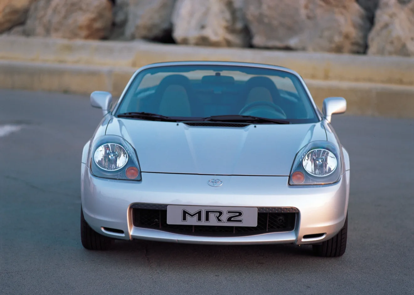 Toyota MR2 prata, visto de frente e com pedras posicionadas ao fundo