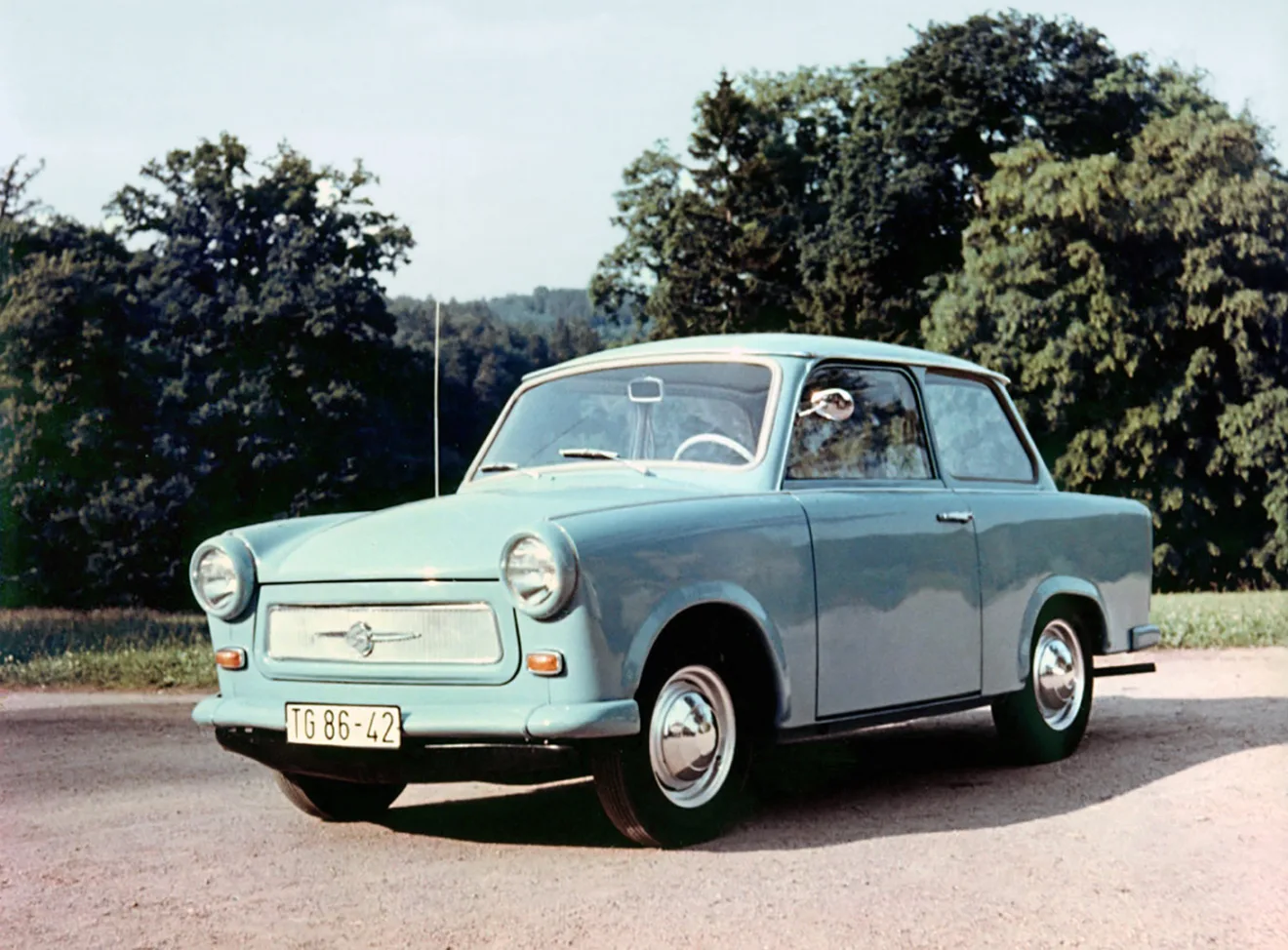 Trabant 601 azul parado de frente no asfalto com árvores ao fundo