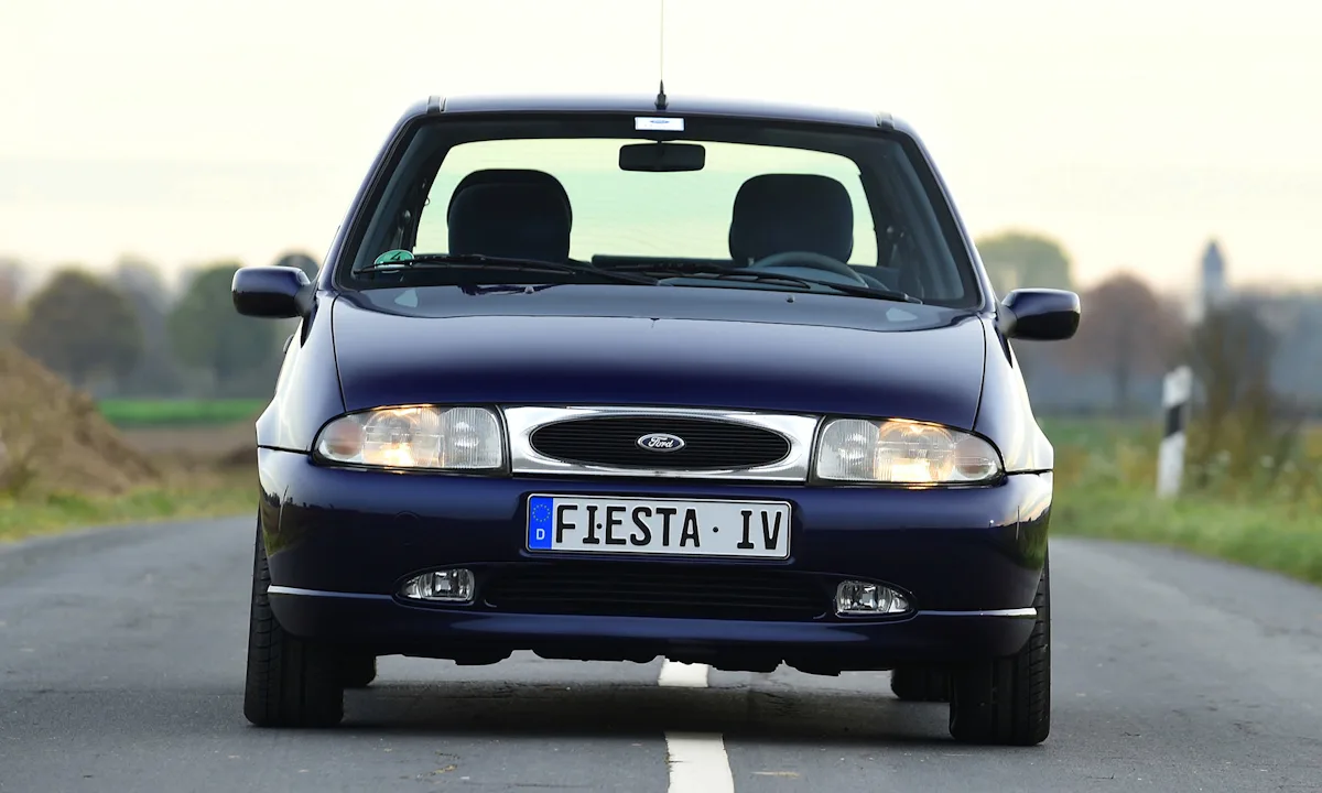 Ford Fiesta tristonho azul com grade frontal cromada visto de frente com os faróis acesos no meio da rua