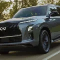 Infiniti QX80 cinza, visto de frente, com faróis acesos e árvores ao redor
