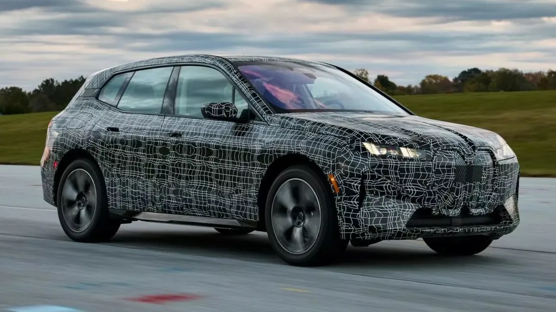 Novo BMW iX reestilizado camuflado de preto e branco, visto de frente, com faróis acesos e andando em uma pista