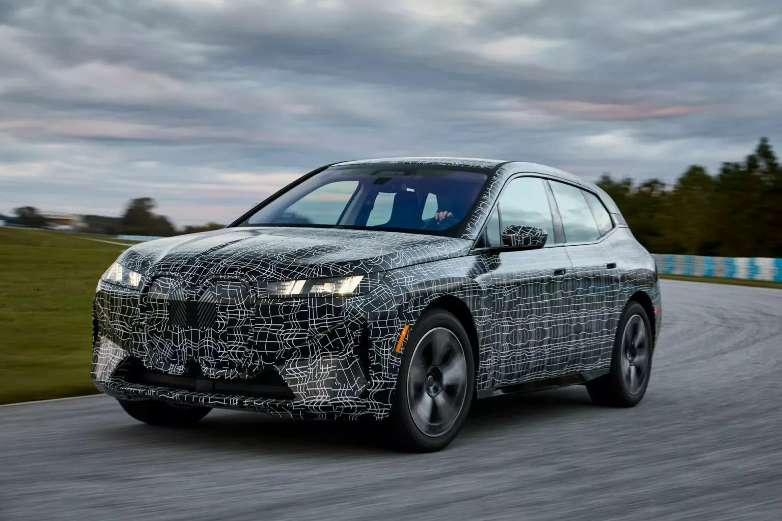 Novo BMW iX reestilizado camuflado de preto e branco, visto de frente, com faróis acesos e andando em uma pista