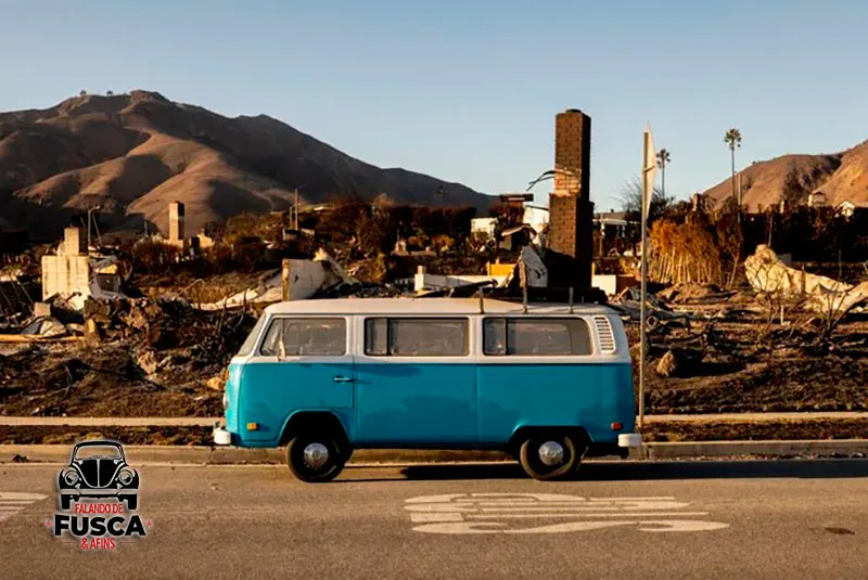 Volkswagen Kombi azul com teto branco, vista de lado e com casas destruídas ao fundo