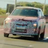 Novo carro barato da Fiat com camuflagem branca e preta, visto de frente, com faróis acesos e placa ao fundo