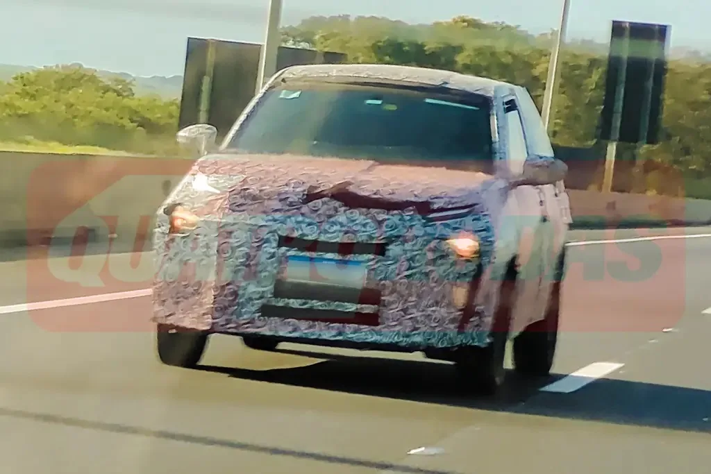 Novo carro barato da Fiat com camuflagem branca e preta, visto de frente, com faróis acesos e placa ao fundo