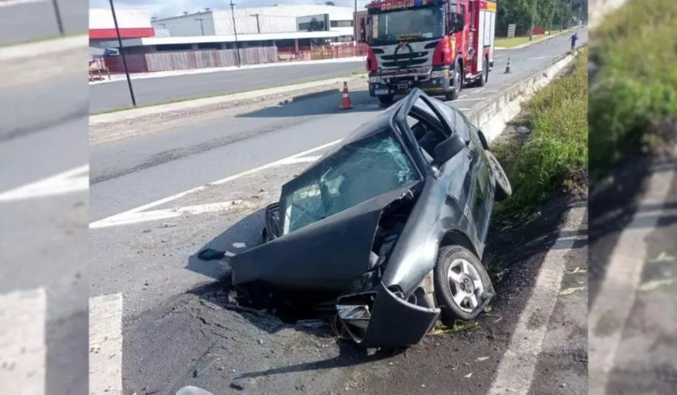 Carro escuro todo destruído após acidente na BR-470 e caminhão dos bombeiros ao fundo