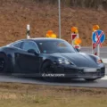 Porsche 718 Cayman elétrico camuflado de preto, visto lateralmente com placas ao lado e faróis acesos