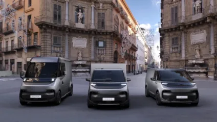 Três vans elétricas da Renault e Volvo, vistas de frente, com prédio ao fundo e DRL's acesos