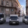 Três vans elétricas da Renault e Volvo, vistas de frente, com prédio ao fundo e DRL's acesos