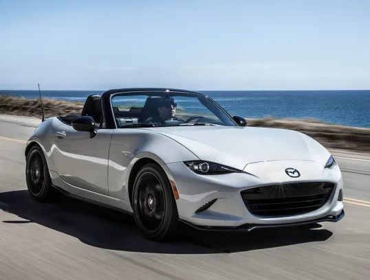 Roadster Mazda MX-5 Miata prata andando na estrada com mar ao fundo