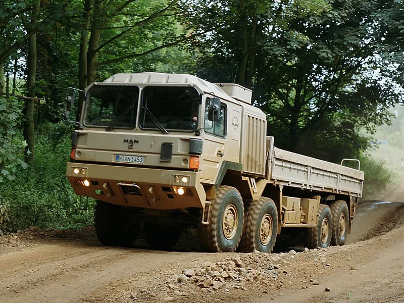 Caminhão Man HX 32.440 BB 8x8 bege, visto de frente e com faróis acesos