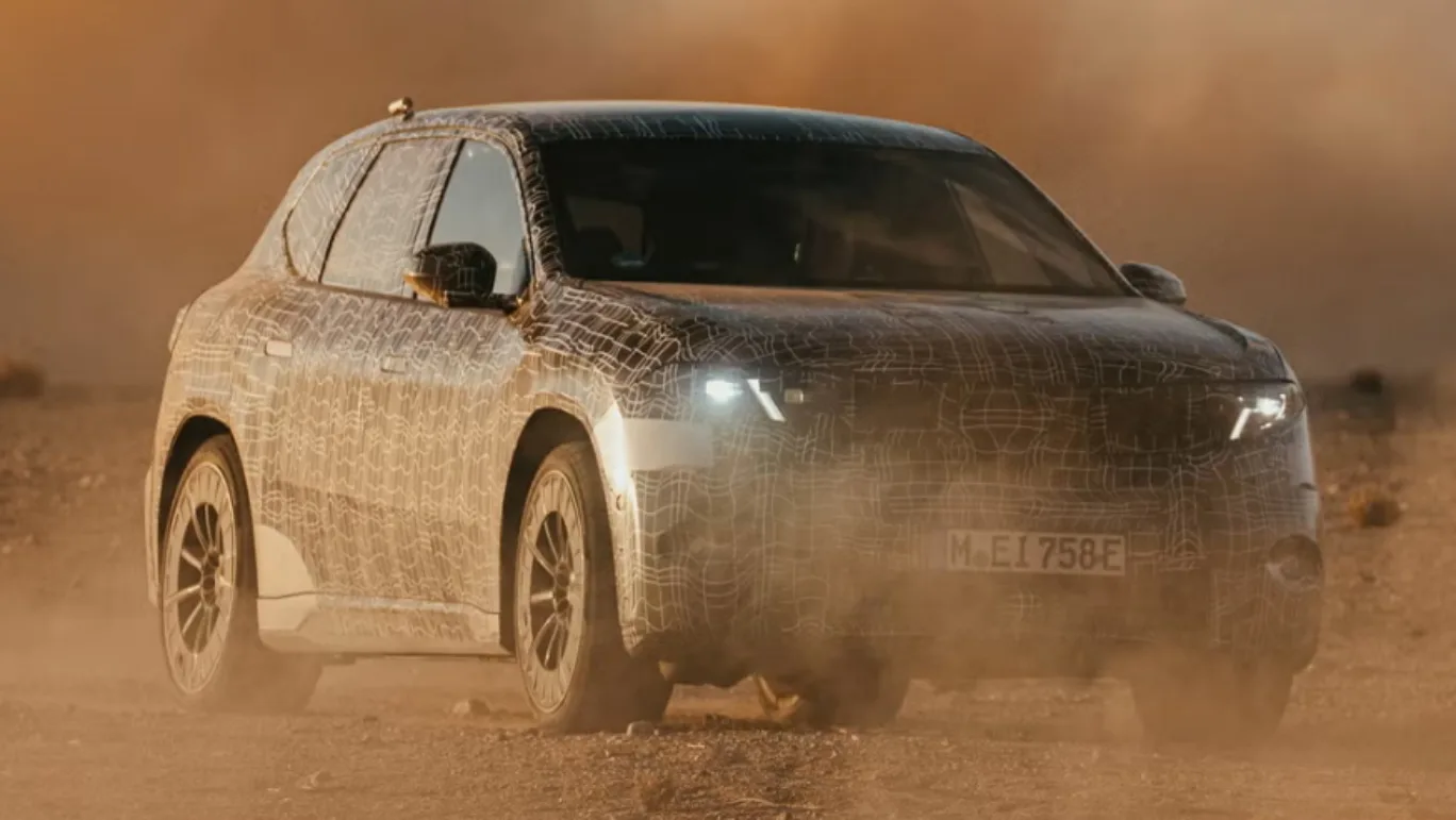 Protótipo camuflado de preto do BMW iX3 Neue Klasse, visto de frente e com faróis acesos