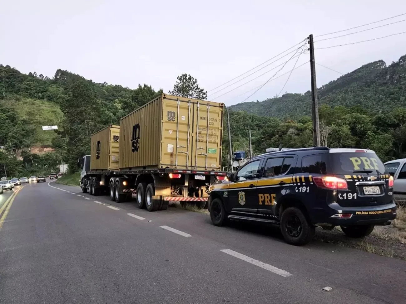 Carreta parada, vista de trás, com carga dourada e viatura da PRF preta parada em seguida