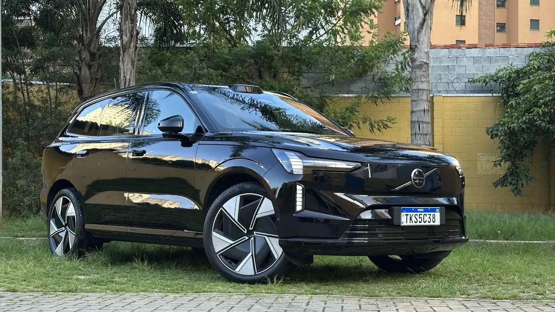 Volvo EX90 Ultra preto parado de frente com muro amarelo e árvores ao fundo Volvo EX90 Ultra preto parado de frente com muro amarelo e árvores ao fundo
